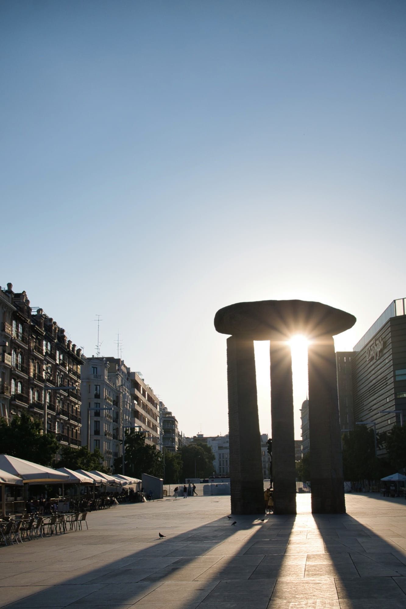Urban plaza at sunrise or sunset with dark trilithon sculpture and starburst sun