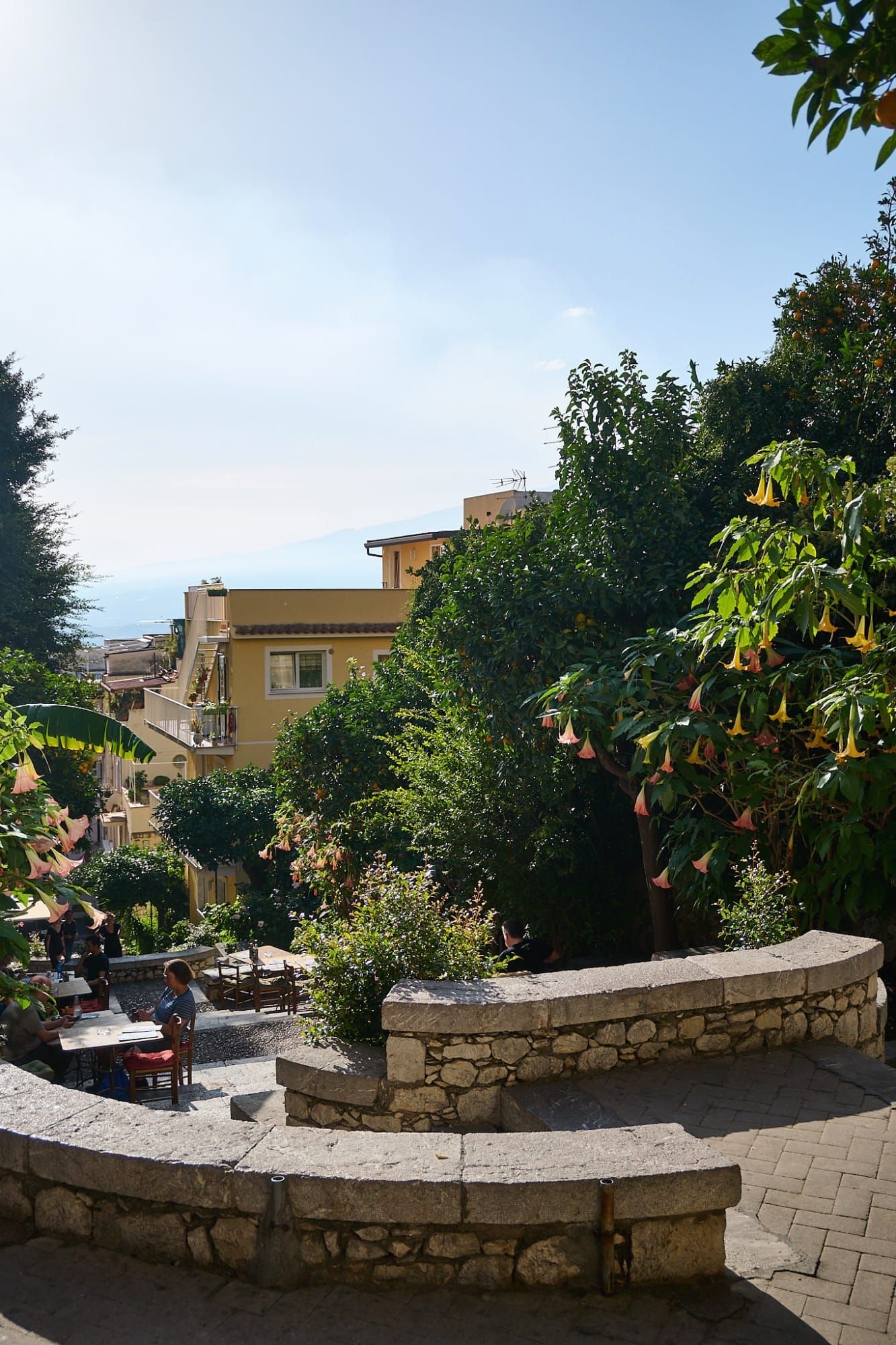 Sunlit terraced restaurant with diners, green trees and yellow buildings overlooking sea