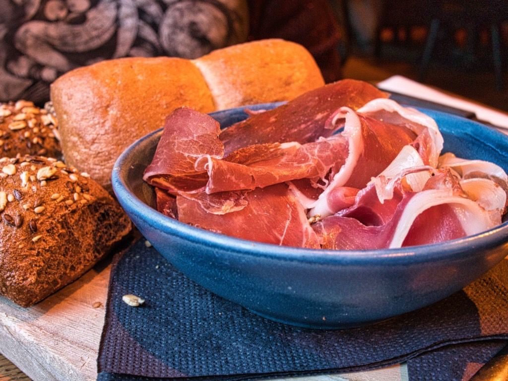 Blue bowl with thin slices of cured ham beside assorted bread on a wooden board