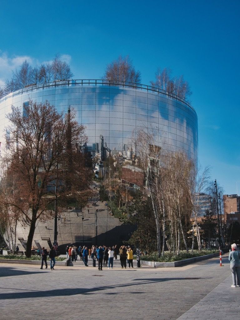 Mirrored cylindrical building reflecting a city square and visitors on a sunny day