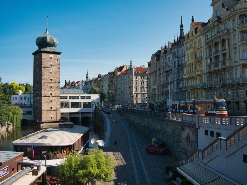 Prague riverside street with old architecture, tram tracks, and a stone tower beside the water