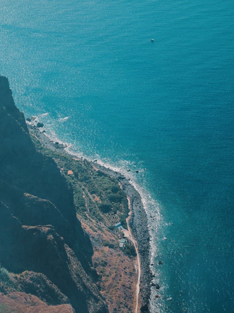 Steep Madeira cliffside dropping to a pebble beach, with a narrow path, a few small houses and bright turquoise Atlantic water