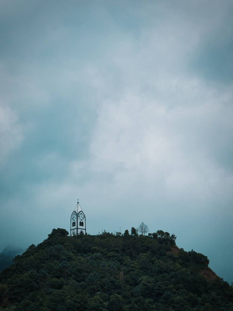 White chapel tower topped with a cross on a forest-covered Madeiran hilltop under a heavy grey sky