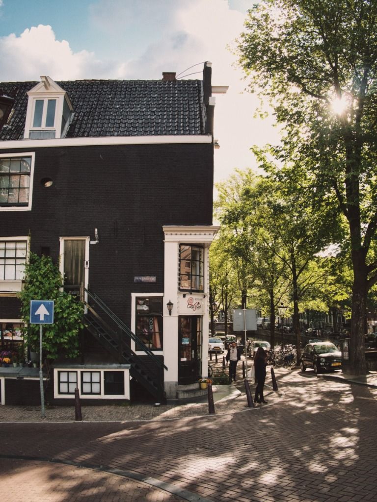 A striking black corner building in Amsterdam with an outdoor staircase, next to a sunlit tree-lined canal with a couple of people walking in the dappled sunlight