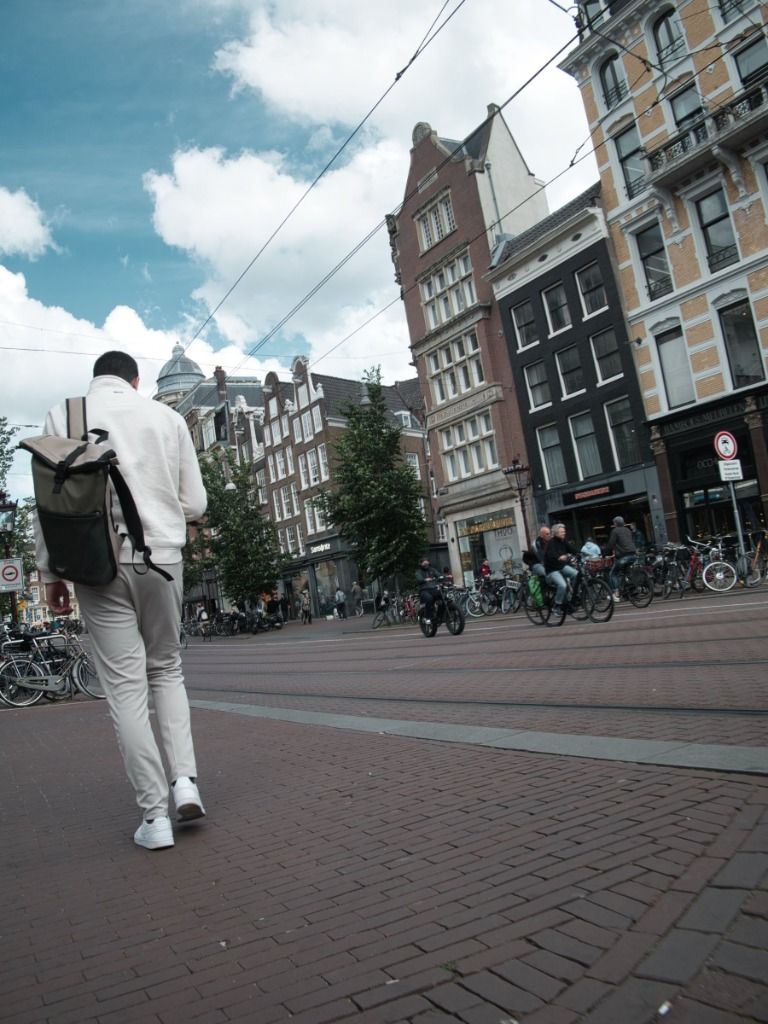 A person with a backpack walking down a brick-paved Amsterdam street, with classic tall Dutch buildings, overhead tram cables, and bicycles in the background under a cloudy sky