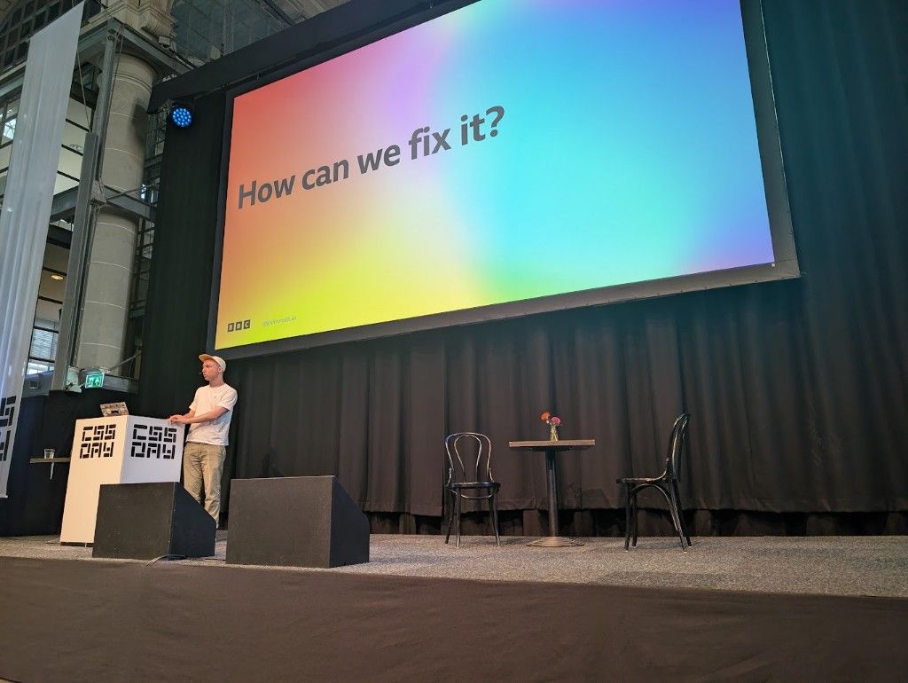 Josh Tumath on stage at CSS Day, with a large projected slide reading 'How can we fix it?' on a colorful gradient background